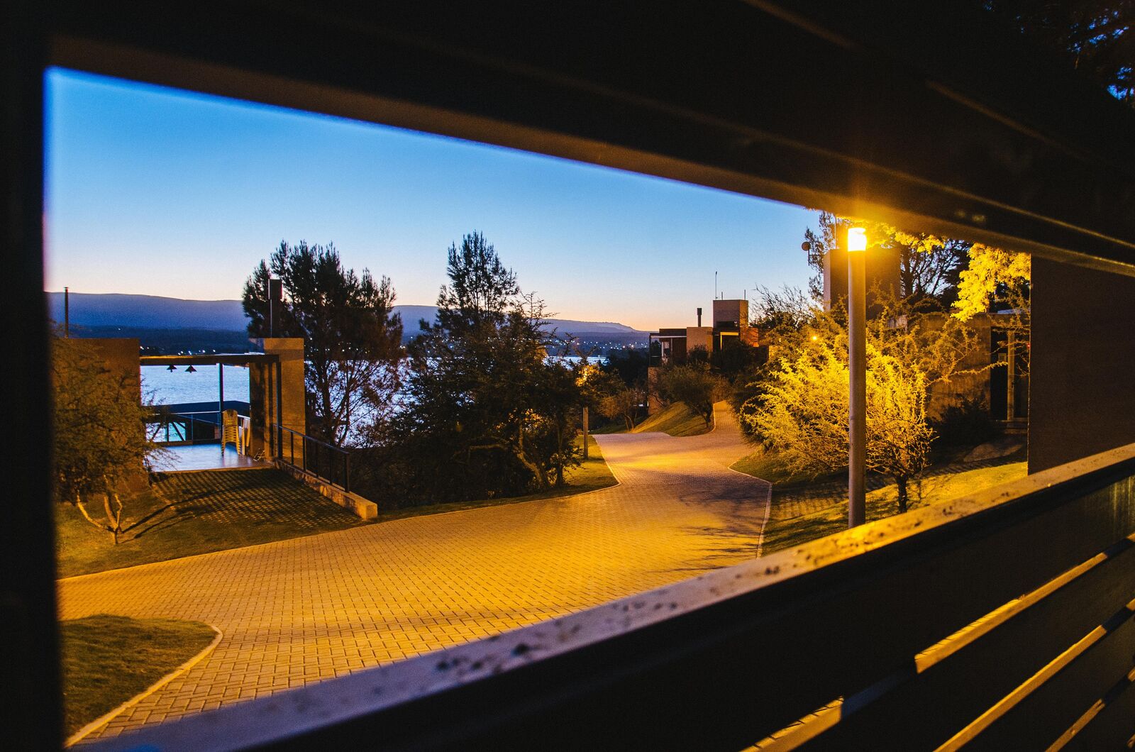 Vista noturna do caminho de acesso iluminado, com o Lago Los Molinos e as luzes do povoado ao fundo