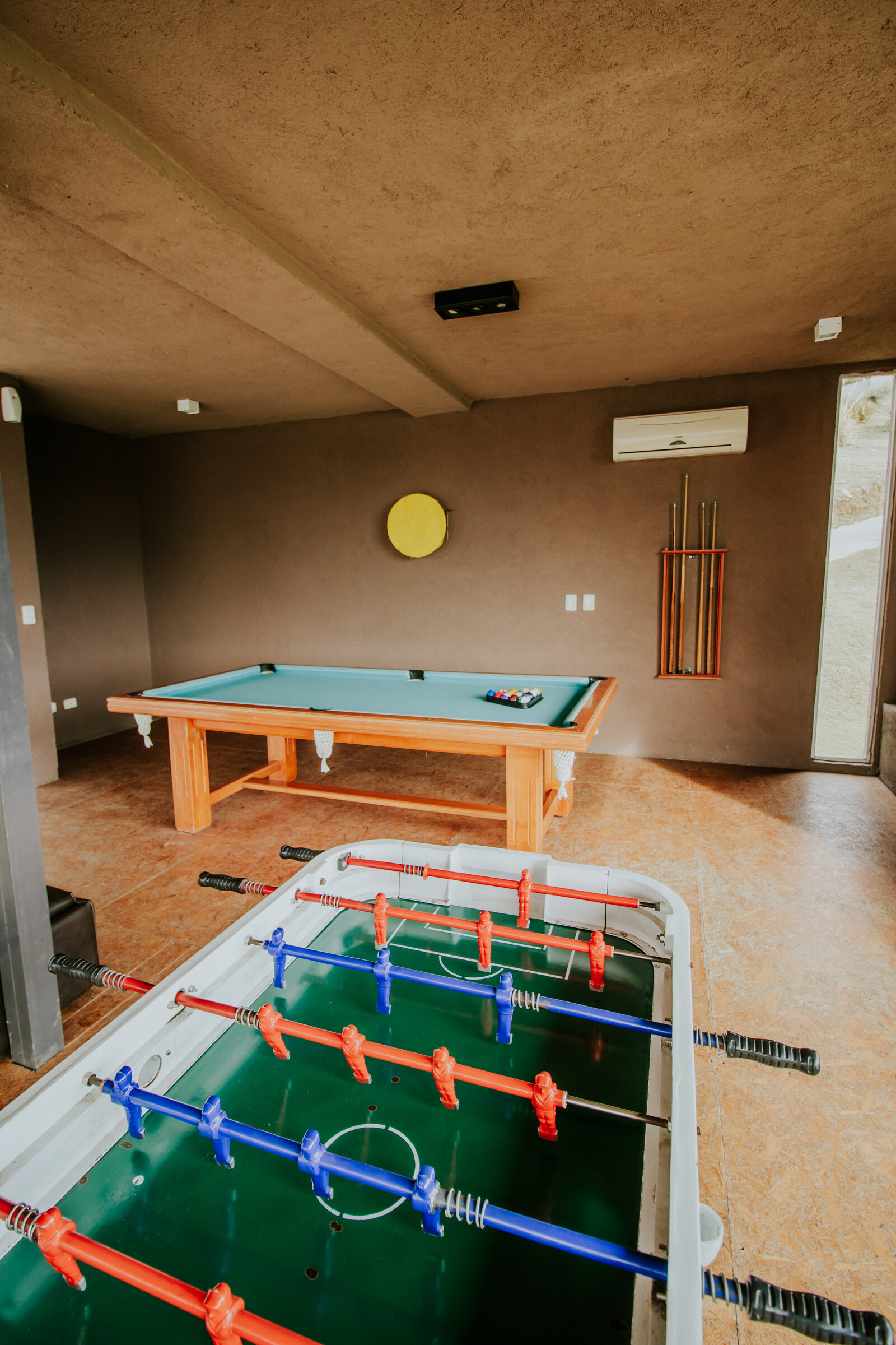 Mesa de bilhar com os tacos na parede e pebolim na sala de jogos
